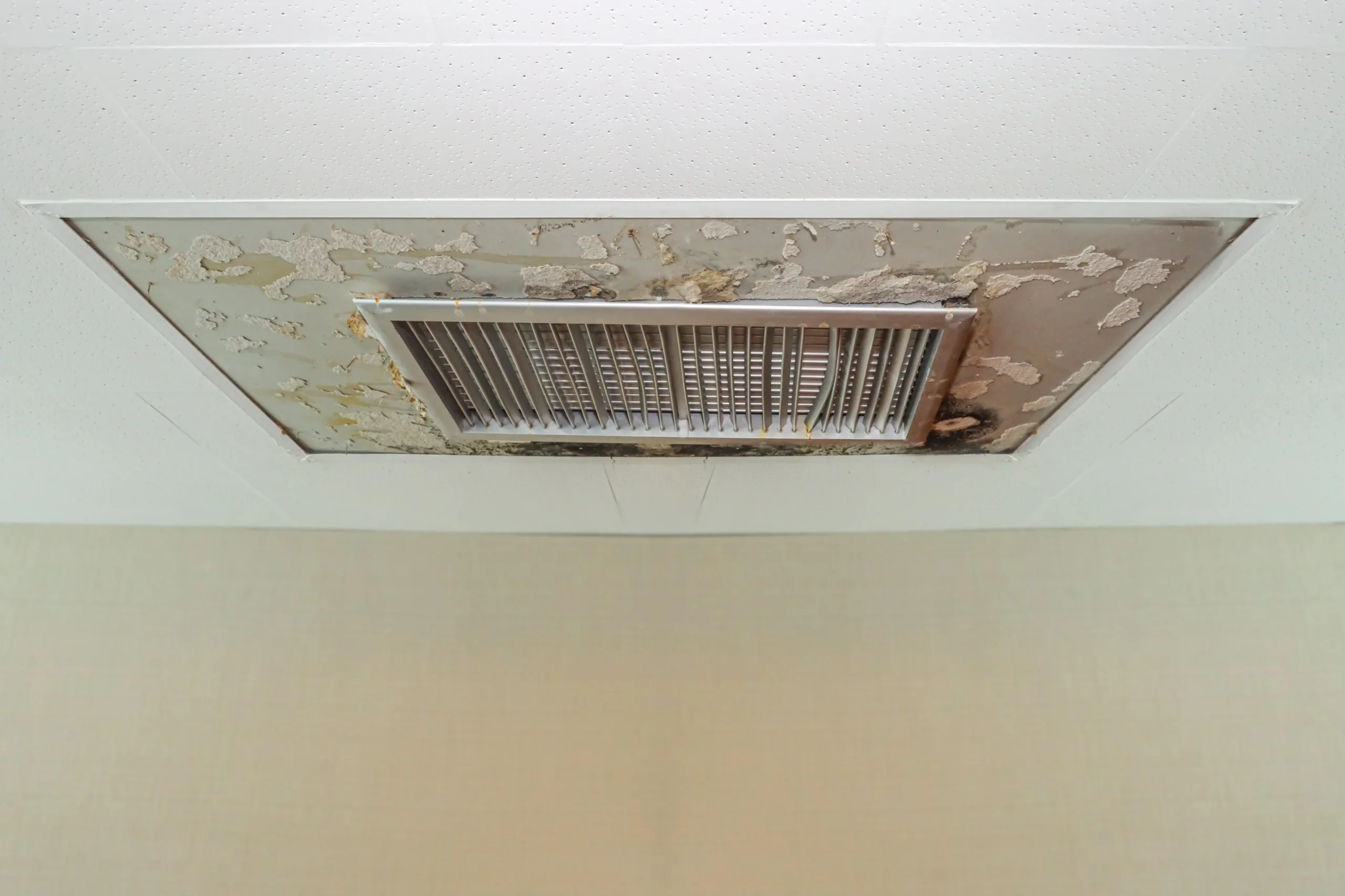 Close-up of a damaged ceiling vent or air conditioning register showing peeling paint, water staining, and possible mold or soot damage around the metal grille.
