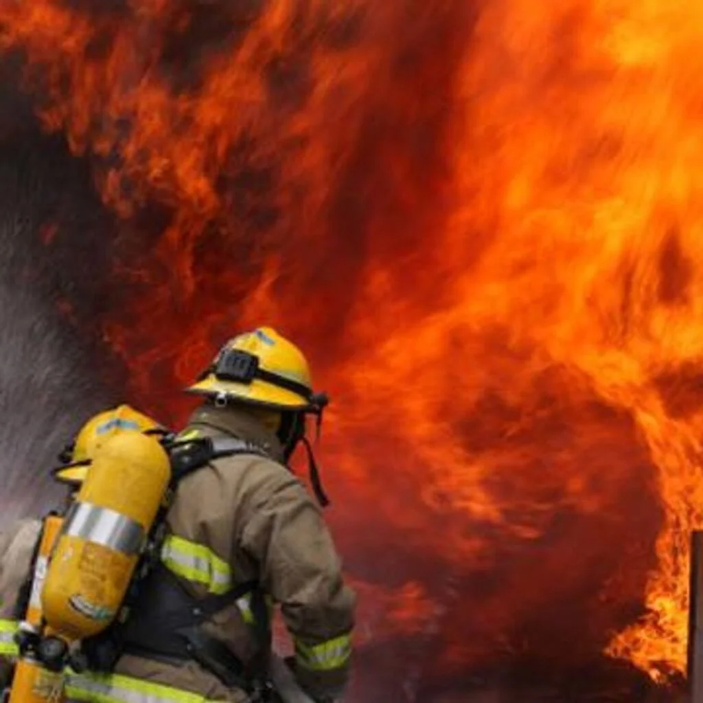 Close-up of two firefighters, wearing yellow helmets and oxygen tanks, facing and combating massive, raging orange and red flames.